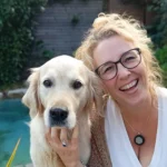 Frau mit blonden Haaren und Brille lächelt ins Bild. Sie umarmt einen weissen Hund.