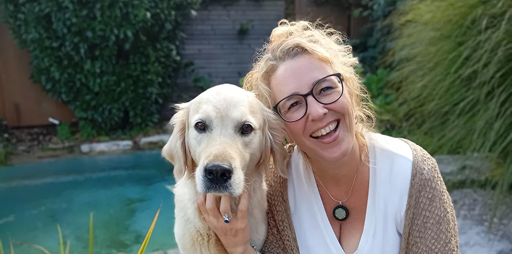 Frau mit blonden Haaren und Brille lächelt ins Bild. Sie umarmt einen weissen Hund.